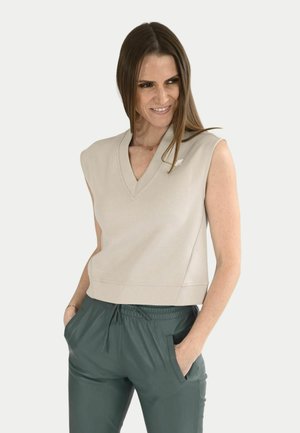 Femme aux cheveux bruns raides portant un top beige sans manches à col en V et un pantalon vert foncé avec cordon de serrage, mains dans les poches.