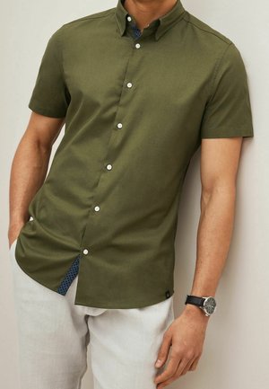 Homme portant une chemise olive à manches courtes boutonnée et un pantalon clair avec une montre noire au poignet gauche.