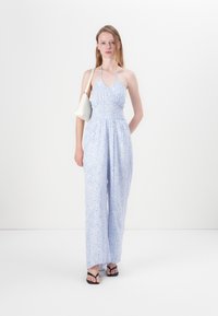 Lys blå blomstrete jumpsuit med halterneck, tettsittende overdel og vide bukser. Brukt med en liten hvit håndveske og svarte sandaler.