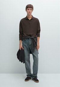 Bruine lange mouwen polo met ribbelmanchetten, gecombineerd met vervaagde blauwe spijkerbroek. Een donkere trui is om de taille geknoopt. Bruine suède schoenen maken de look compleet.