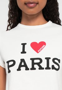 Femeie îmbrăcată într-un tricou alb cu textul "I [inimă roșie cu FUTURE] PARIS" pe față, arătând o parte din fața și partea superioară a torsului.