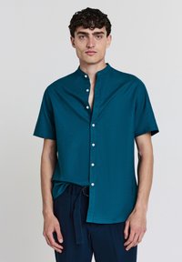 Türkises Kurzarm-Hemd mit Stehkragen, verfügt über eine glatte Textur und weiße Knöpfe entlang der Knopfleiste. Getragen mit dunklen Hosen.