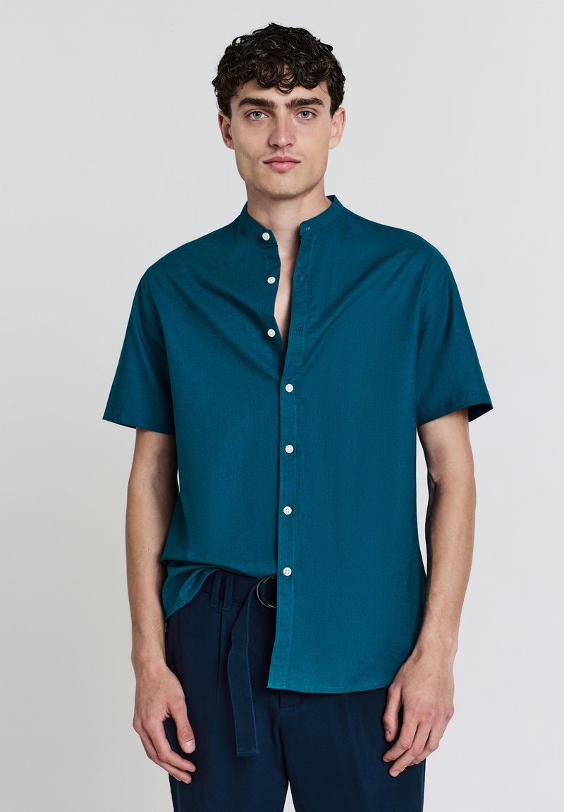 Türkises Kurzarm-Hemd mit Stehkragen, verfügt über eine glatte Textur und weiße Knöpfe entlang der Knopfleiste. Getragen mit dunklen Hosen.