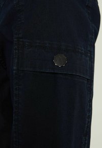 Dark blue fabric with a soft texture; features a side pocket, secured with a black snap button with a subtle logo design.