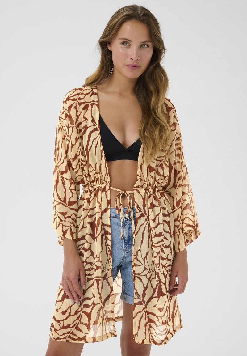 Kimono estampado en naranja y beige, con cinturilla ajustable. Presenta mangas largas y una tela ligera y semitransparente. Llevado sobre pantalones cortos y un bikini.