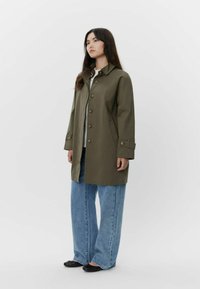 Cappotto verde oliva con collettino a punta, grandi bottoni e tasche laterali, indossato con jeans larghi in denim blu. Tessuto liscio con una vestibilità strutturata.