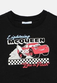 Fekete pulóver, amelyen egy vörös rajzfilmautó látható kék szemekkel és fehér lángokkal. A szöveg vastag betűkkel "Lightning McQueen" és "Like a Flash" feliratokat tartalmaz.