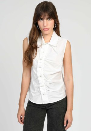 Jeune femme aux longs cheveux bruns portant des boucles d'oreilles argentées en forme de créoles, une chemise blanche sans manches froncée avec boutons, et un pantalon gris foncé, fond neutre.