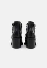 Botines de cuero negro hasta el tobillo con cremallera lateral y correa ajustable. Presentan un tacón robusto y detalles de costura para agregar textura.