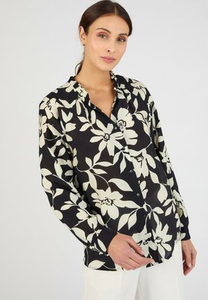 Femme portant un chemisier noir à boutons avec de grandes imprimés floraux blancs et un pantalon blanc, posant devant un fond clair uni.