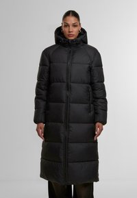 Lång svart puffjacka med quiltade sektioner, hög krage och dragkedja fram. Har sidfickor och en huva med dragsnören.
