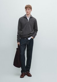 Tmavě šedý svetr na zip, pod ním bílá košile s límečkem, tmavé džíny, hnědé kožené boty, držící burgundskou tašku.