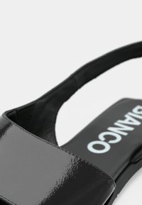 Sandalia de charol negro con una amplia tira y diseño de punta abierta. La plantilla tiene texto blanco sobre fondo negro.