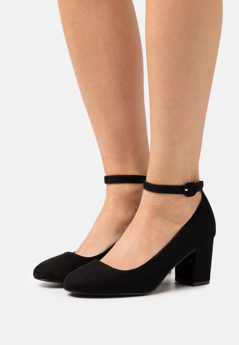 Jambes portant des chaussures en daim noir à talons blocs avec des brides à la cheville et des bouts ronds, debout sur un fond clair uni.