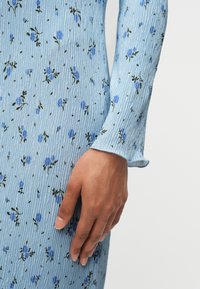 Κοντινό πλάνο ενός χεριού που ακουμπάει σε ένα φόρεμα από υφή υφάσματος ανοιχτού γαλάζιου χρώματος με μικρά μπλε λουλουδένια σχέδια και μακριά μανίκια.