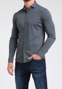 Camiseta gris de manga larga hecha de tela suave con cuello clásico, diseño delantero con botones y silueta ajustada. Combinada con jeans de mezclilla azul.