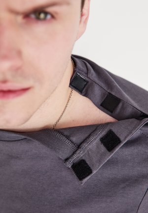 Szara koszulka z miękką fakturą, z zapięciem na rzepy przy kołnierzu i okrągłym dekoltem. Zbliżenie na detal przy ramieniu.