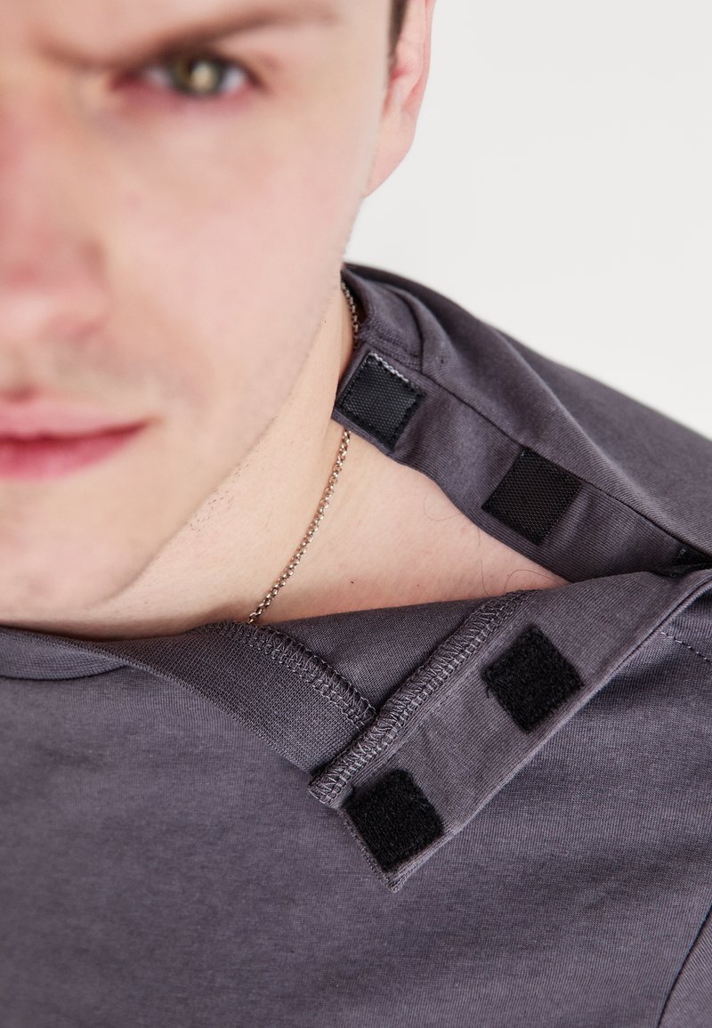 Γκρί μπλούζα με μαλακή υφή, διαθέτει κλείσιμο με βέλκρο στον γιακά και στρογγυλή λαιμόκοψη. Κοντινή λήψη της λεπτομέρειας του ώμου.