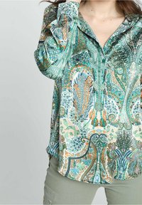 Femme portant une blouse à manches longues avec un motif cachemire détaillé en vert, bleu et orange, accompagnée de pantalons vert clair.