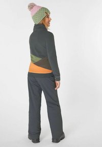 La giacca in pile presenta un design a blocchi di colore con grigio scuro, verde, marrone e arancione. Indossata con pantaloni scuri e un berretto lavorato a maglia a motivi.