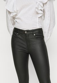 Calças skinny de imitação de couro preto com cintura alta, bolsos frontais e fecho com botão metálico; textura suave e design ajustado.