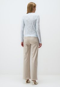 Top lungo in pizzo blu chiaro a maniche lunghe con motivi floreali abbinato a pantaloni beige a gamba larga, indossato con tacchi di colore crema.