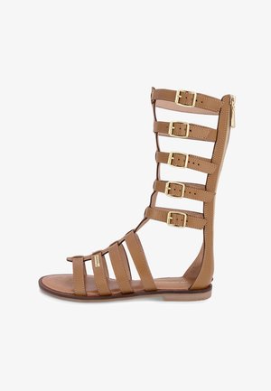 Sandali gladiatore marroni con molteplici cinturini regolabili e dettagli in metallo dorato. Materiale in pelle liscia con suola piatta.