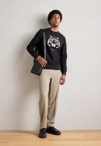 Schwarzer Pullover mit einem Tigermotiv, kombiniert mit beigen Hosen. Das Model hält eine kleine schwarze Umhängetasche und trägt schwarze Slippers.