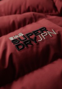 Section d'une veste rembourrée rouge avec un grand texte brodé noir et blanc indiquant "SUPER DRY JPN" sur un tissu lisse au fini mat.