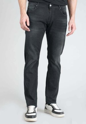 Jean en denim noir avec une coupe fuselée, présentant des poches avant, une fermeture éclair et une texture légèrement délavée. Associés à des baskets noires et blanches.