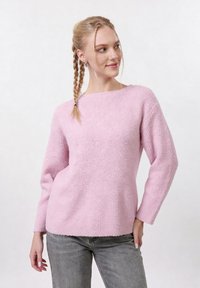 Jeune femme aux cheveux tressés portant un pull duveteux rose et un jean gris, posant les mains détendues sur un fond clair uni.