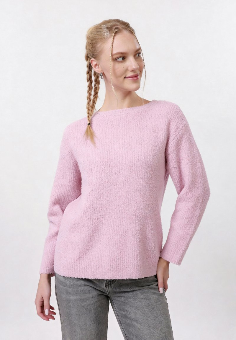 Jeune femme aux cheveux tressés portant un pull duveteux rose et un jean gris, posant les mains détendues sur un fond clair uni.