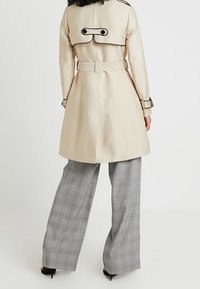 Beżowy trencz z czarnymi dodatkami, z paskiem w talii i dwurzędowym zapięciem. W komplecie szare, szerokie spodnie w kratę.