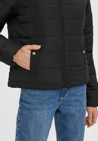 Sort polstret jakke med lynlås foran, med sidelommer og synlige sømme. Sammen med blå denim jeans.