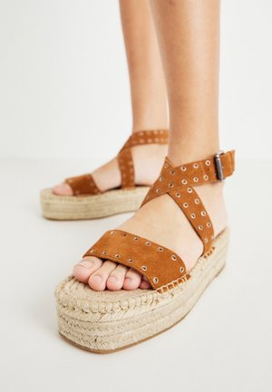 Brown suede platform sandals with crossed straps, metal eyelets, and a jute-wrapped wedge sole. Adjustable ankle strap for fit.