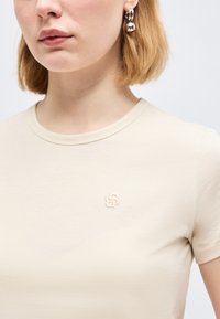 Γυναίκα που φοράει μπεζ T-shirt με στρογγυλή λαιμόκοψη, με μικρό κεντητό λογότυπο στο στήθος και ασημένια σκουλαρίκια κρίκους με κρεμαστές σφαίρες.