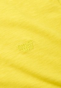 Gelbes Baumwollgewebe mit einer dezenten Textur und einem gestickten "Superdry"-Logo. Das Design ist schlicht und ungeschmückt.