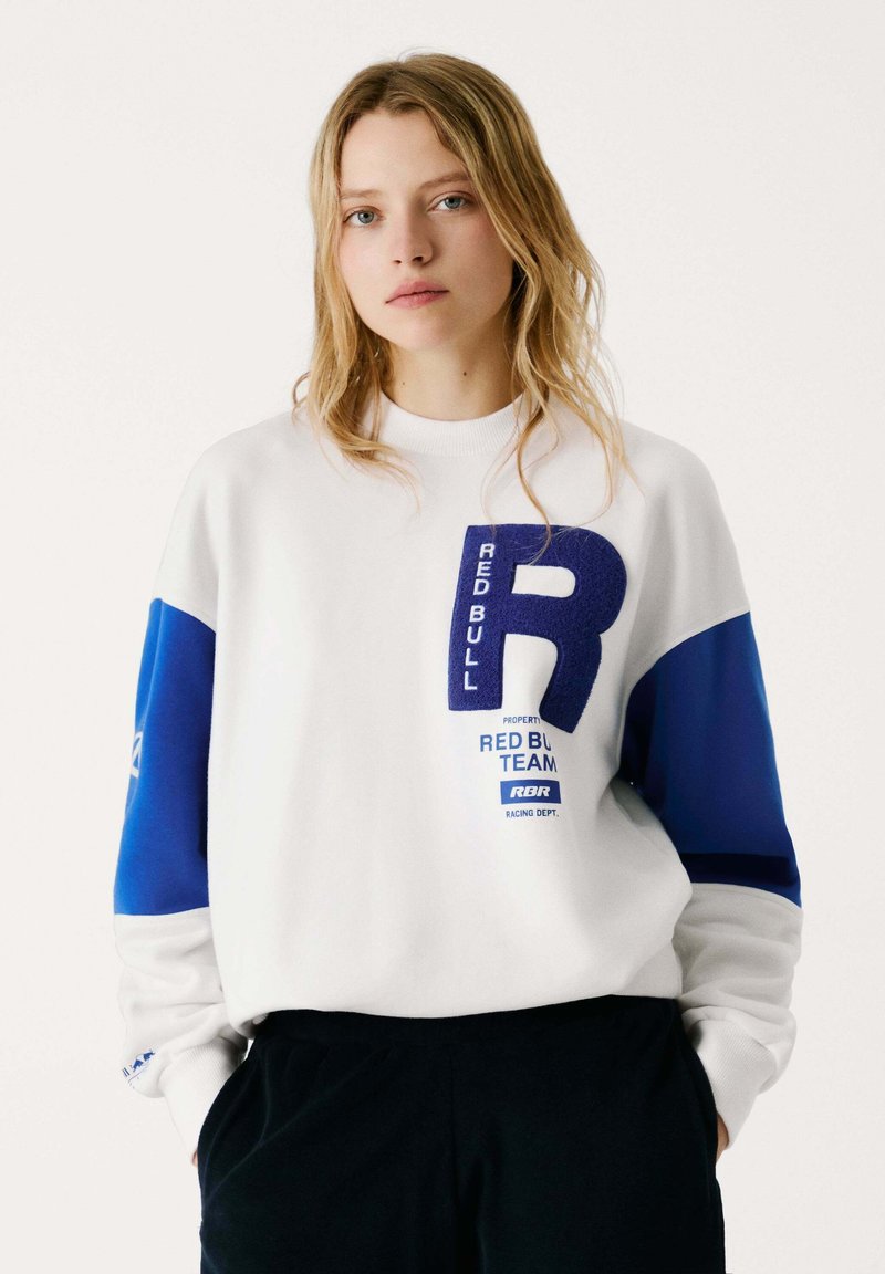 Felpa bianca con maniche blu, caratterizzata da un grande logo blu a "R" e dal testo "RED BULL TEAM". Il materiale sembra liscio e informale.