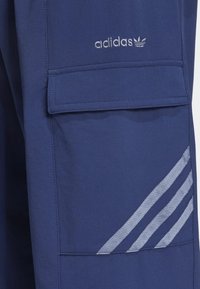 Námořnické modré cargo kalhoty s hladkou texturou, obsahující štítek na kapsu a tři reflexní stříbrné pruhy na boku. Logo Adidas ve stříbrné barvě.