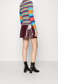 Pull rayé à manches longues en orange, bleu et vert, associé à une jupe bordeaux brillante. Bottines noires en vernis avec un talon carré.