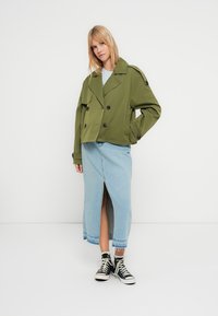 Oliivinvihreä lyhyt takki, jossa on kaksirivinen napitus, yhdistetty vaaleansiniseen farkkuhameeseen, jossa on etuhalkio, sekä mustat tennarit.