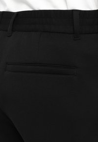 Pantalon noir sur mesure avec détail de poche passepoilée à l’arrière et passants de ceinture à la taille, présenté sur fond blanc.