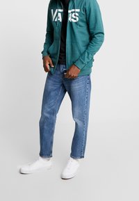 Felpa zip con cappuccio color turchese con grande logo bianco, abbinata a una maglietta nera e jeans in denim azzurro chiaro. Sneakers bianche completano l'outfit.