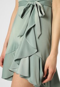 Mintgrön omslagsskjorta gjord av lättviktsmaterial, med en flödande volangkant och ett matchande knytband i midjan. Slät textur.