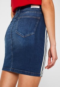 Falda de mezclilla en azul oscuro con un diseño ajustado, que presenta dos franjas blancas laterales y bolsillos traseros. Dobladillo limpio y detalles mínimos.