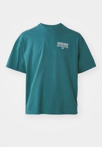 Teal cotton t-shirt with a crew neck, short sleeves, and a printed graphic that includes "VERT'ERE BERLIN" on the front.
