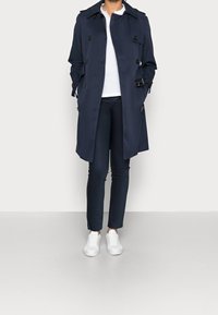 Navy trench coat with double-breasted design, large buttons, and shoulder epaulets, paired with fitted navy pants and white sneakers.