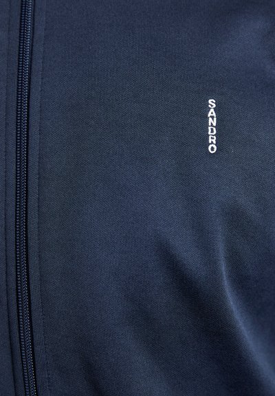 Marinblå zip-up-jacka i slät tyg, med en strukturerad yta och en liten vit "SANDRO"-logga nära dragkedjan.