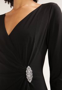 Kvinna klädd i en svart V-ringad klänning med långa ärmar, samlad vid midjan med en dekorativ silver- och pärlbroche.