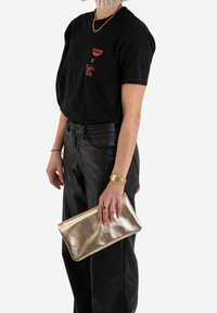 Metaliczne złote kopertówka trzymana w ręce, o gładkiej fakturze. Czarny t-shirt z czerwonymi grafikami i czarne skórzane spodnie dopełniają strój.